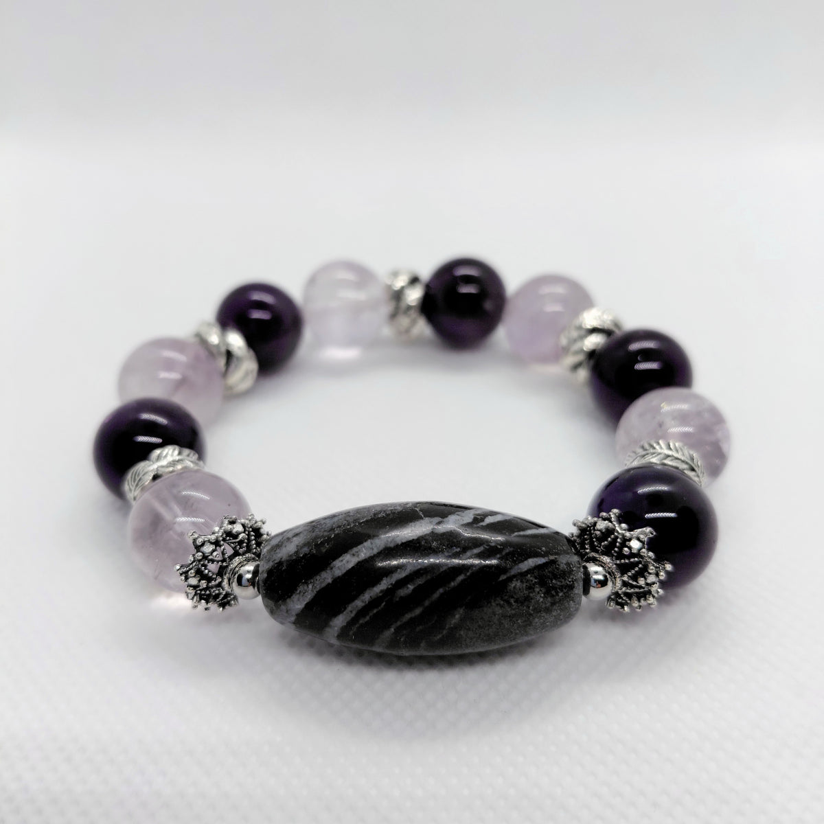 A beaded stretch bracelet featuring dark purple amethyst beads, light lavender crystal beads, a large black and white striped teardrop bead, and silver-toned metal spacers, displayed on a white textured background.